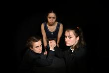 Three woman, two of them clasping hands as in an arm-wrestling match, with black background