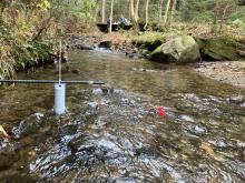 A sensor placed in a Vermont forested stream with students collecting data in the background.