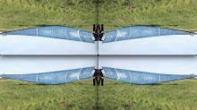 A digitally manipulated image divided into four mirrored quadrants. The top half of the image is an upside-down mirror of the bottom half, and the left side mirrors the right, creating a symmetrical, kaleidoscope-like effect. The primary image shows a person dressed in black, standing on green grass, holding the edge of a large, translucent blue piece of fabric with white markings that stretches out horizontally.