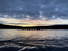 An 8-shell of rowers cuts across a serene Lake Dunmore with the setting sun visible in the background.