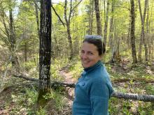 a woman with brown hair pulled back into a bun smiles at the camera. She is wearing a teal sweater and is standing in the woods.