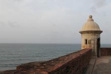 A part of the wall and a white stone turret of the fort in Old San Juan stands in the foreground with the Atlantic Ocean behind it.