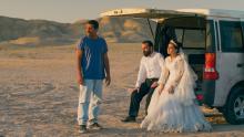 Three people are watching a sunset in the middle of a desert. Two of them sit in the open trunk of a truck and are dressed as the groom and the bride.