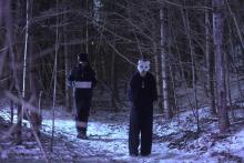 Two people in snowy woods, one wearing a mask and facing the camera, one facing away from the camera
