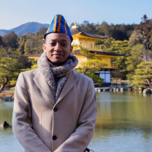 Male with hat on by pond with building behind.