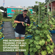 Smiling student wearing tan ball cap, black shirt and blue shorts, holding cucumbers, standing in a raised bed garden.