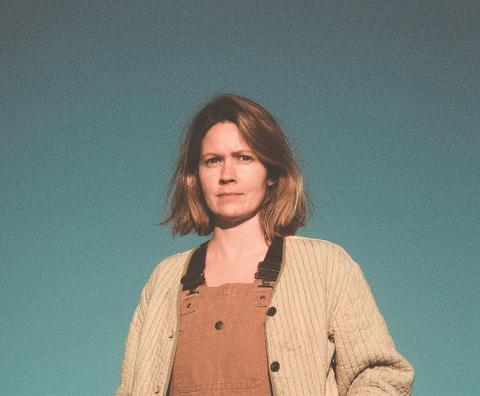 person in brown overalls and a sweater standing in front of a green background