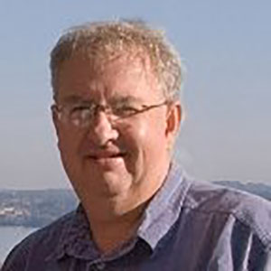 Dr. Charles Colgan, in the sun with a peek of water (a bay) behind him, he is sort of smiling and squinting in the sun