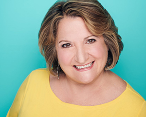 Eleanor Kerlow smiling brightly, posed headshot style in a yellow blouse against a turquise background