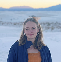 Elle Bent with a slight smile, with beach, dunes, mountains at sunset in the background