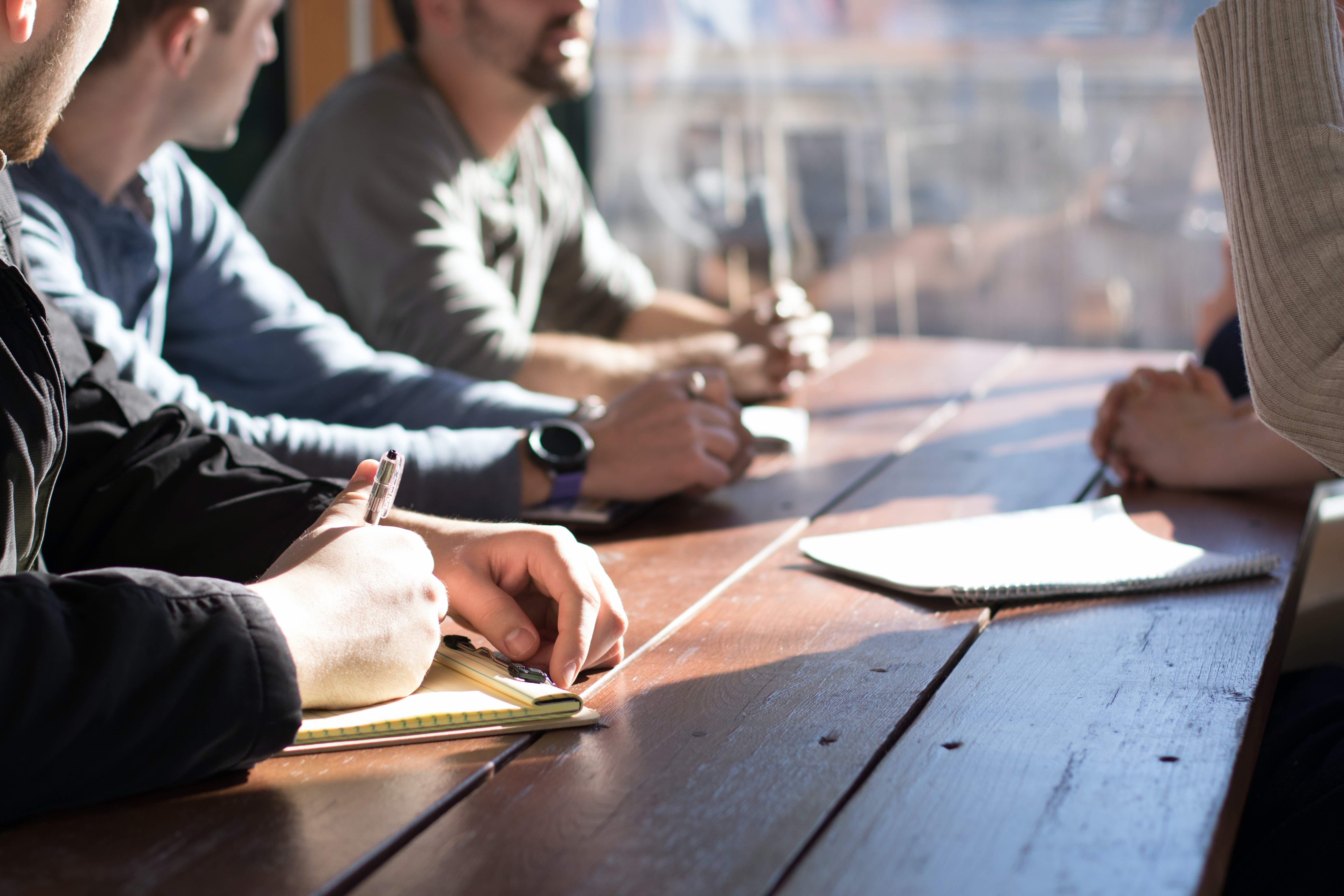 Image of 4 adults at a wooden conference table, only showing their hands, as they take notes as if in a meeting