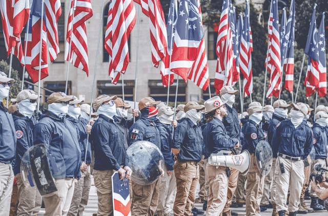 individuals stand wearing hats and faces covered, holding American flags