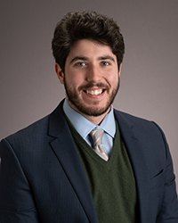 Jack Anderson, a young man with a full smile, bright eyes, and dark hair, mustach and trim beard, wearing a tie and business suit