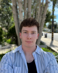 Nate looking pensive in a blue striped shirt against a green and tree-lined street