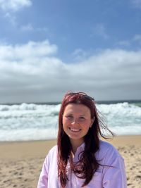 Elena smiling with the wind in her red hair, on the beach with the white capped waves breaking behind and blue sky above