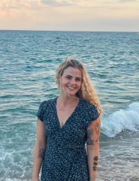 Sophie smiling in a blue dress staning on the beach in front of a blue ocean