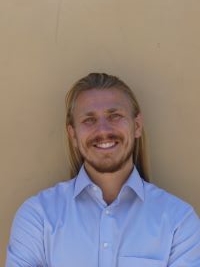 Thomas smiling brightly and sporting blue colored shirt, long hair, beard and mustache