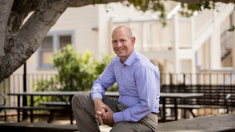 Jason Blazakis sitting on a bench outside under a tree smiling at the camera