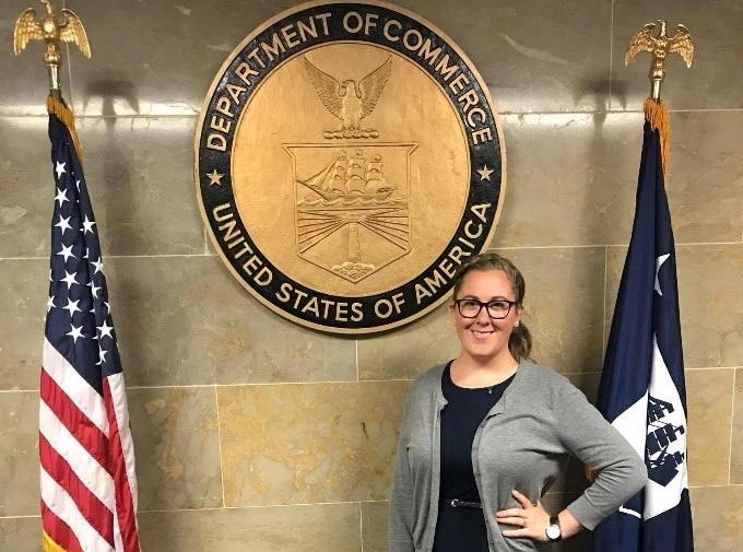 Woman standing in front of the Department of Commerce seal on a wall flanked by the US flag and another flag.