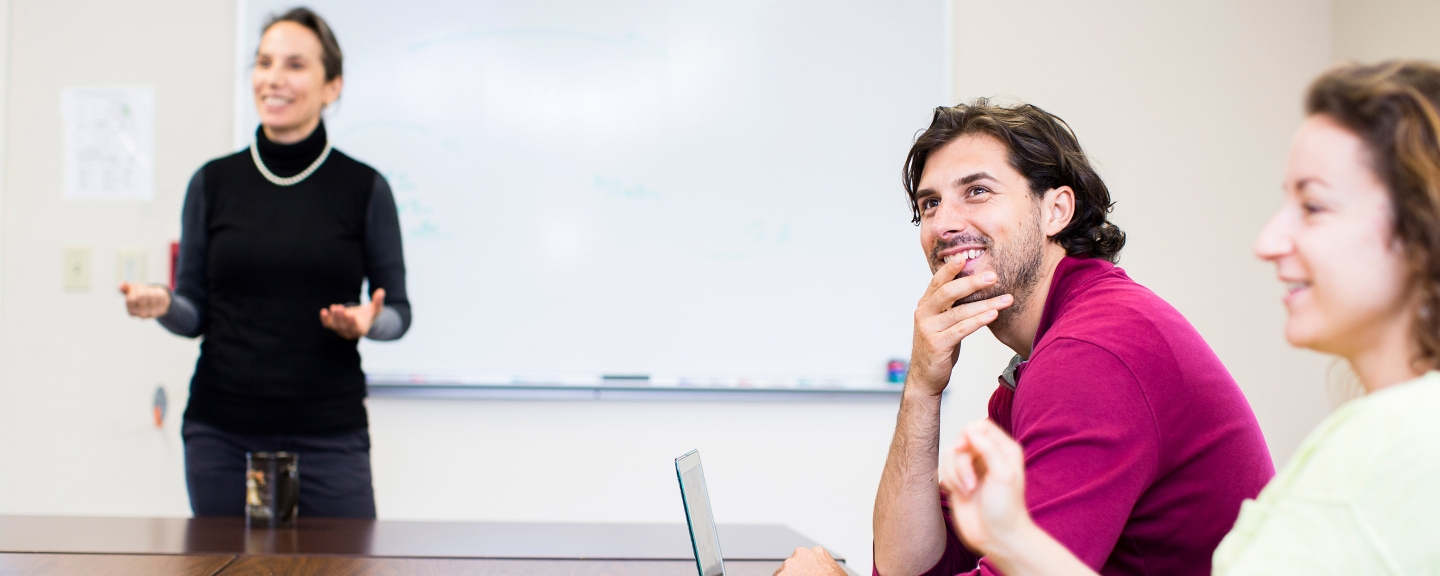 Students in a classroom smiling and listening to a professor.