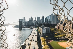 The skyline of Manhattan as seen through a cut our chain link fence--cars driving, baseball field, skyscrapers bridge and water all can be seen