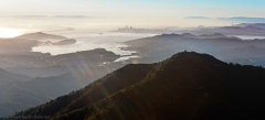 A view of San Francisco Bay taken from the north, looking south toward the SF skyline, coastal hills in foreground, sunlight gleaming off the water, leading to the SF skyline that mixes ephemerally with the clouds behind