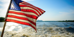 a flag flys off the stern of a vessle, with ocean and city scape in background