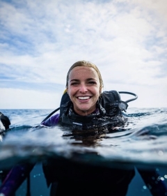 Whitney Berry, in a dive suit, smiling brightly with head above water in the blue ocean, a blue sky with white clouds behind