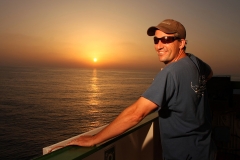 Dr. Brandon Southall, on the bow of a ship with the sun setting over the ocean behind him, looking happy in his T-shirt, ball cap, and sunglasses