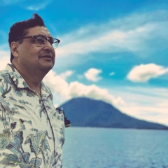 Dr. Adil Najam, dark hair, glasses, Hawaian shirt in profile with blue ocean, island, and sky in background