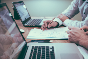 image of two people sitting at desk, just their hands writing on paper in front of their laptop computers