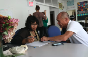 two people sitting at a desk looking at paper materials, one instructing the other