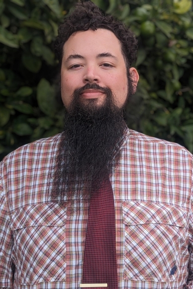 Bernard wearing a plaid shirt and tie, standing in front of green foliage