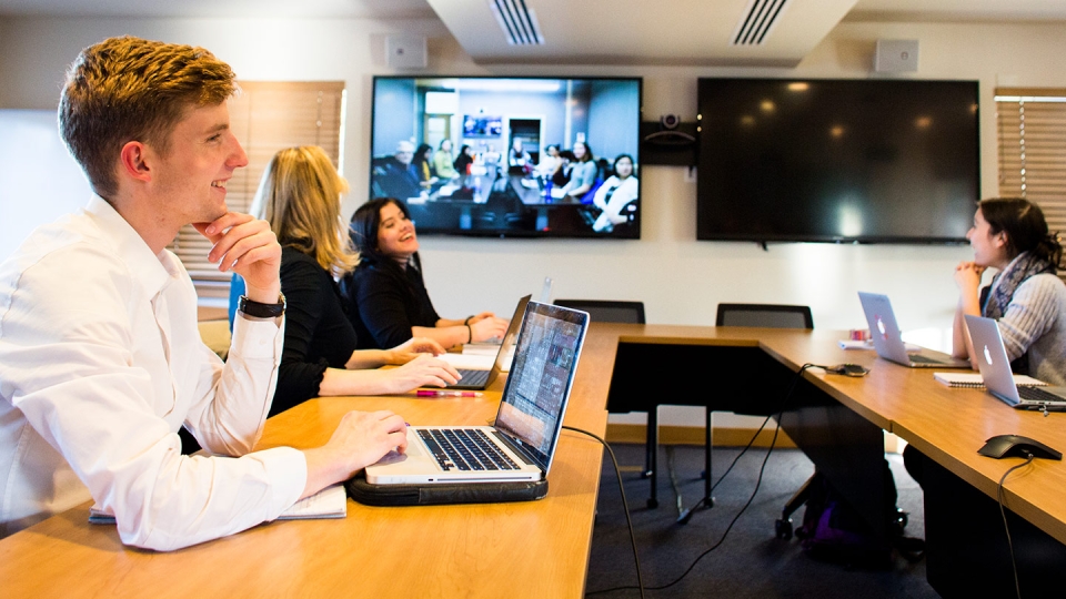 Four students in a classroom video conferencing with other students who appear on a large screen mounted to the wall.