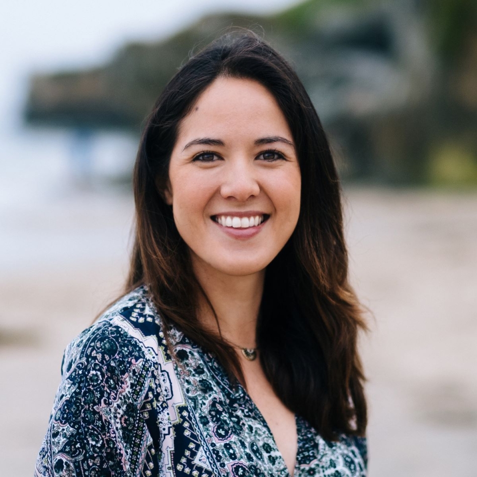 Ms. Mariko Powers, headshot, with a bright smile, dark, long hair, smiling eyes outdoors with a beach and cliff in the background