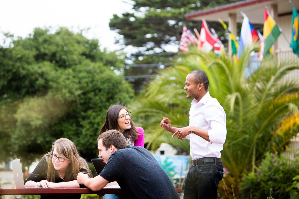 Four students around a table outside. Two are sitting looking at a laptop while their two colleagues stand and talk. Flags and palm trees in the background.