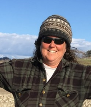 Monique Fountain, Director of the Tidal Wetland Program at the Elkhorn Slough National Estuarine Research Reserve. She is wearing a warm, knit cap and flannel shirt, smiling brightly with red cheeks and sunglasses against a blue sky with clouds, and the greens and browns of the slough.