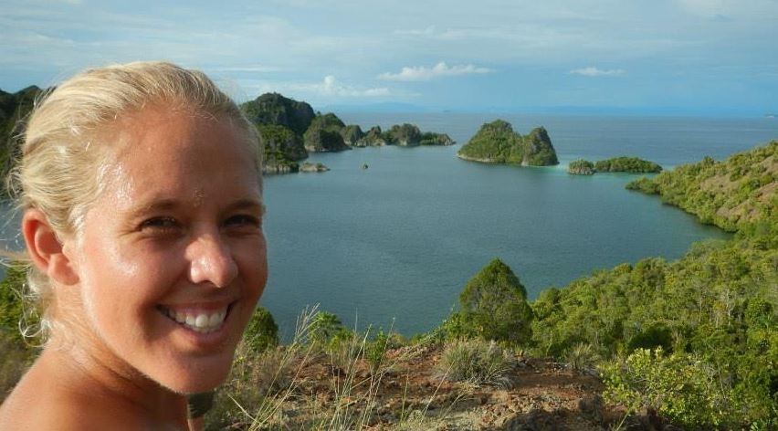Whitney Yadao-Evans smiling and squinting a bit looking into the sun, with an incredibly beautiful scene behind her--an ocean filled Hawaiian caldera, green mountains in a circle, ocean in the middle and on the outside