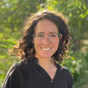 Lindsay Cope smiling and looking confident, glasses, black blouse, curly dark hair, against a verdant green background