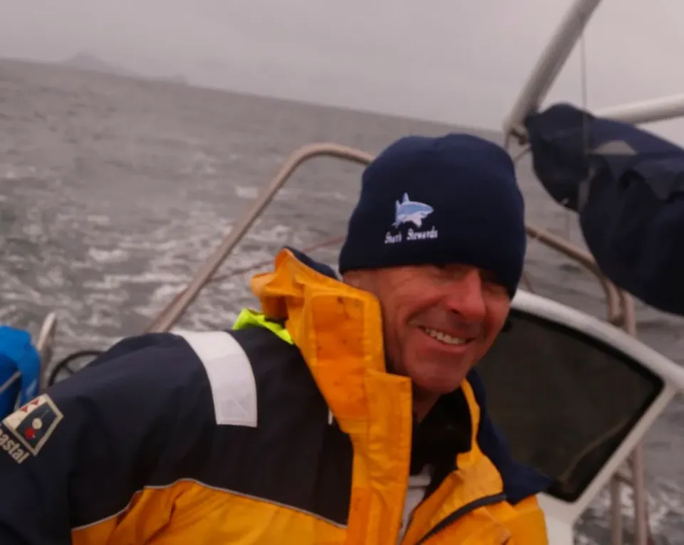 David McGuire riding on a sea-going vessle, wearing an orange coat and black beanie--looks cold and grey, but he looks absolutely happy