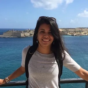 Zara Currimjee against a blue sky and blue water on a sunny day, leaning against a rail, smiling brightly, long black hair, sunglasses on her head