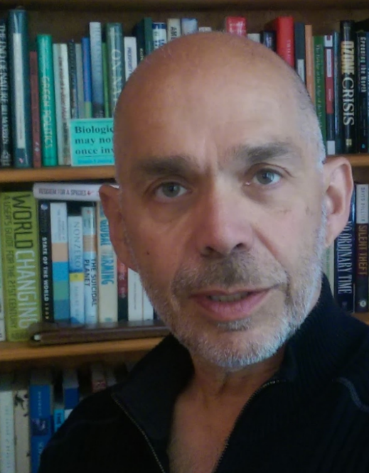 Tom Athanasiou looking straight at camera, earnest and concerned, stubble on his face, black shirt, no hair, against a backgrounnd of books