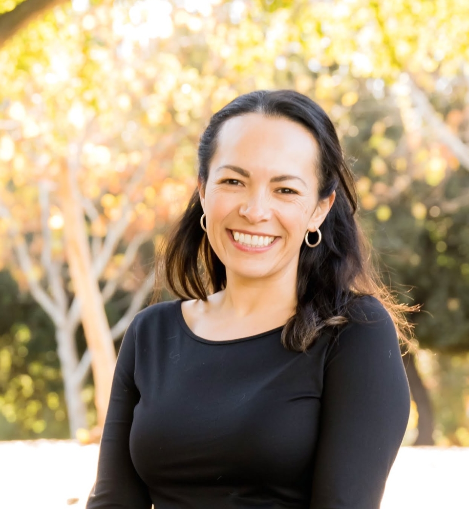 Silvia Sanchez Bor smiling with her whole self, with dark hair and black shirt, against a backdrop fo golden leafed trees