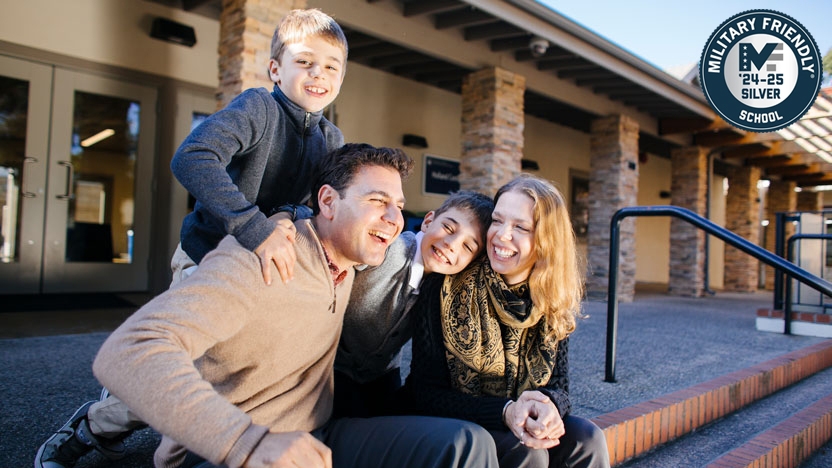 Family of two parents and two children sitting outside a building on campus.