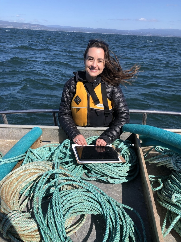 Kacy Cooper on a boat with a big smile, wind in her hair, laptop resting on coiled roaps, wearing a yellow life vest.