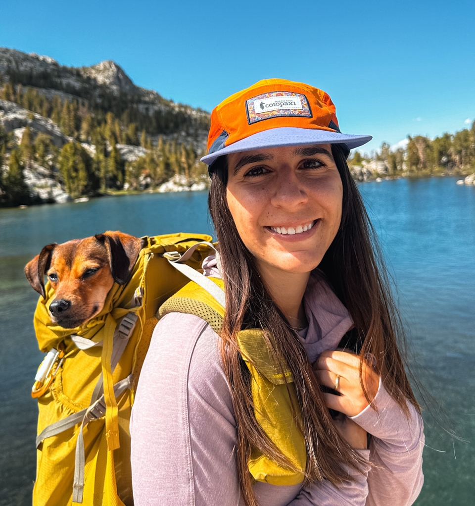 Melis Okter in what must be the best headshot ever, because not only is she looking bright and smiling, but she has the cutest ever doggie in her yellow backpack, and she is standing with the crystal blue waters of a mountain lake behind her.  