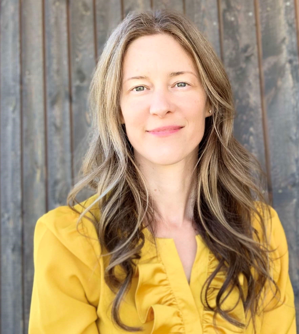 Laura Birkes looking directly at camera with subtle smile, she has long hair, wearing yellow blouse, standing against wooden fence