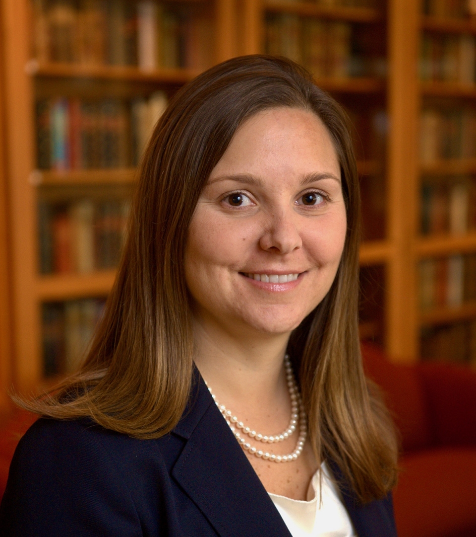 Dr. Joanna Lewis smiling and wearing pearls and business attire, standing in front of bookshelf lined with books