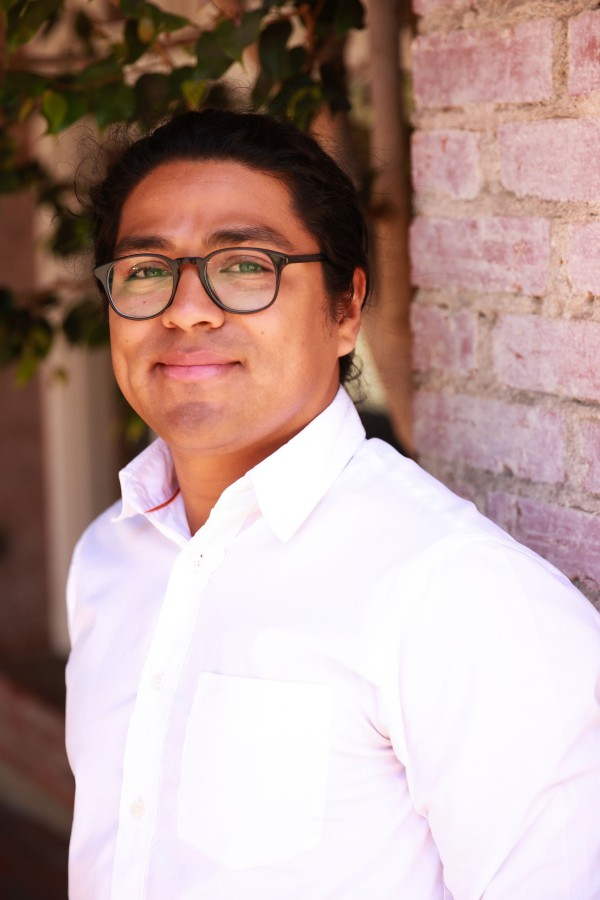 Richard Castillo smiling and looking smart in black glasses and white collared shirt standing against a background of plants and red brick wall