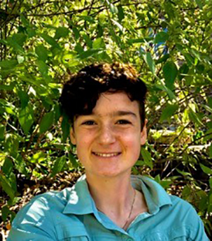 Charley Peebler smiling with a turquoise shirt, short brown hair, she is standing in front a green tree with sunlight streaming through it